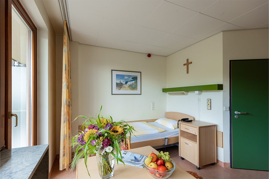Ein besipielhaftes Patientenzimmer mit Bett und Tisch. Auf dem Tisch steht ein Blumenstrauß, eine Obstschale und eine Zeitung.
