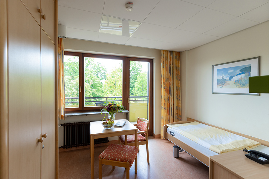 Ein beispielhaftes Patientenzimmer mit Schrank, Bett, Sitzecke und Balkon.