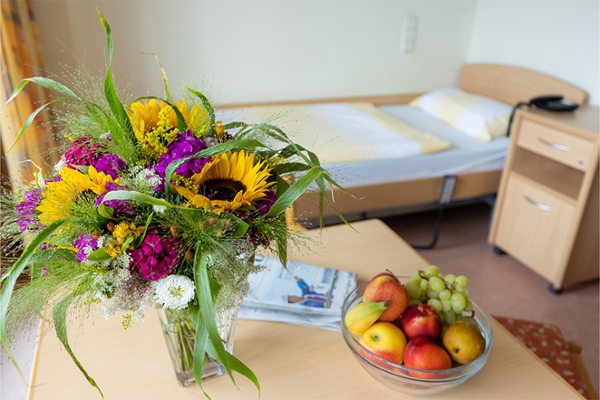 Auf dem Tisch eines Patientenzimmers steht ein Blumenstrauß in einer Vase, ein Schale mit verschiedenem Obst und eine Zeitung. Auf dem Tisch eines Patientenzimmers steht ein Blumenstrauß in einer Vase, ein Schale mit verschiedenem Obst und eine Zeitung.