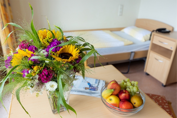 Auf dem Tisch eines Patientenzimmers steht ein Blumenstrauß in einer Vase, ein Schale mit verschiedenem Obst und eine Zeitung.