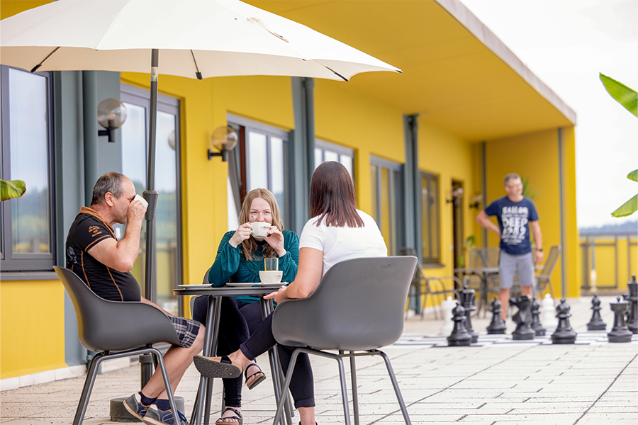 An einem Tisch sitzen mehrer Personen, trinken Kaffee und unterhalten sich. Im Hintergrund spielt ein Mann auf einem Außenschachbrett.